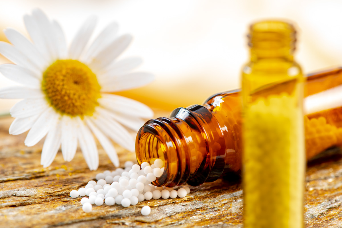 Braune Glasflasche mit weißen homöopathischen Kügelchen auf Holz, weiße Kamillenblüte im Hintergrund